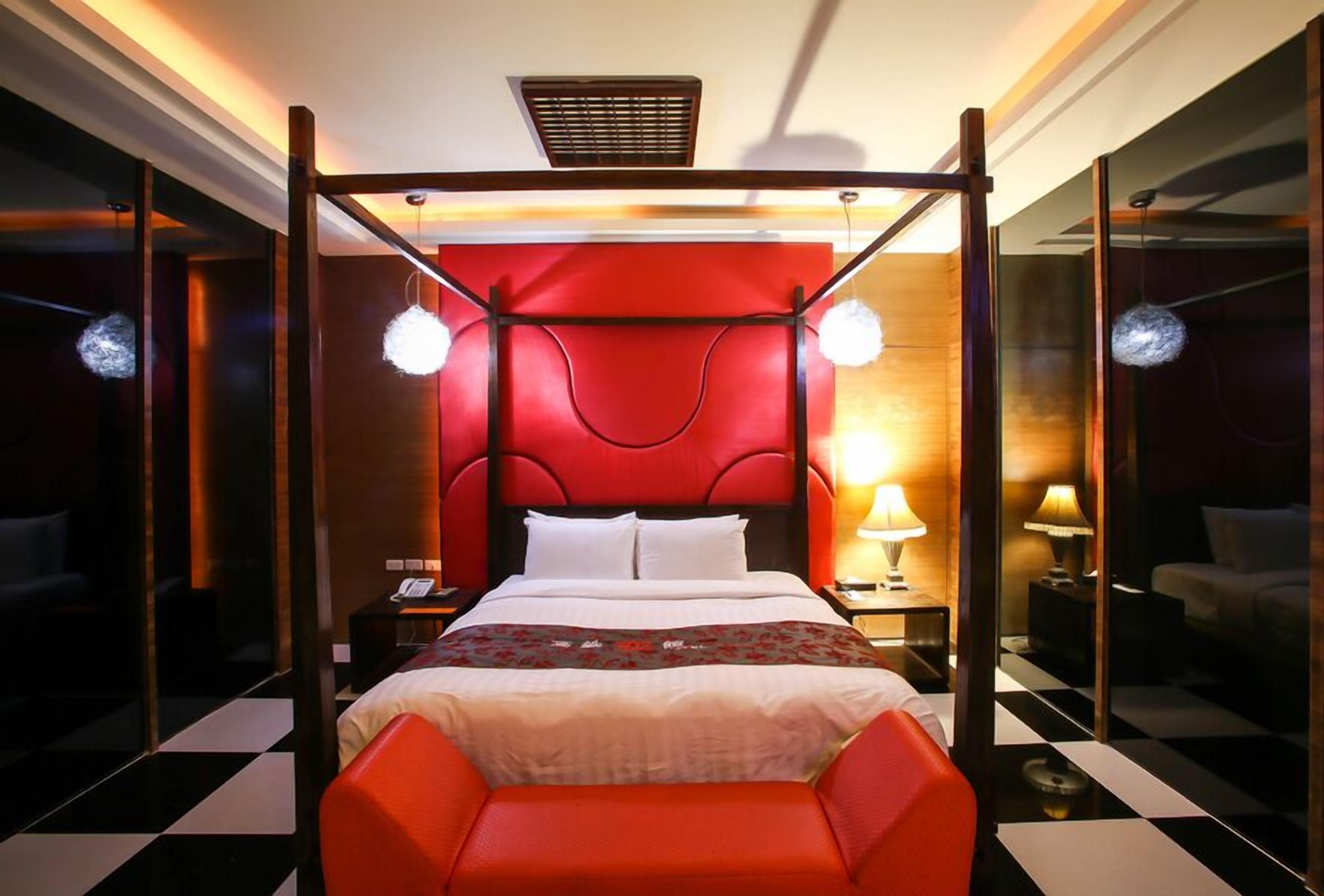 Una habitación de hotel con una cama con dosel, cabecera roja, dos lámparas de noche, mesitas de noche y suelo a cuadros.
