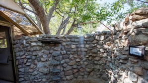 A stone-walled outdoor shower area with a wooden ladder, a metal sign, and a tree providing shade.