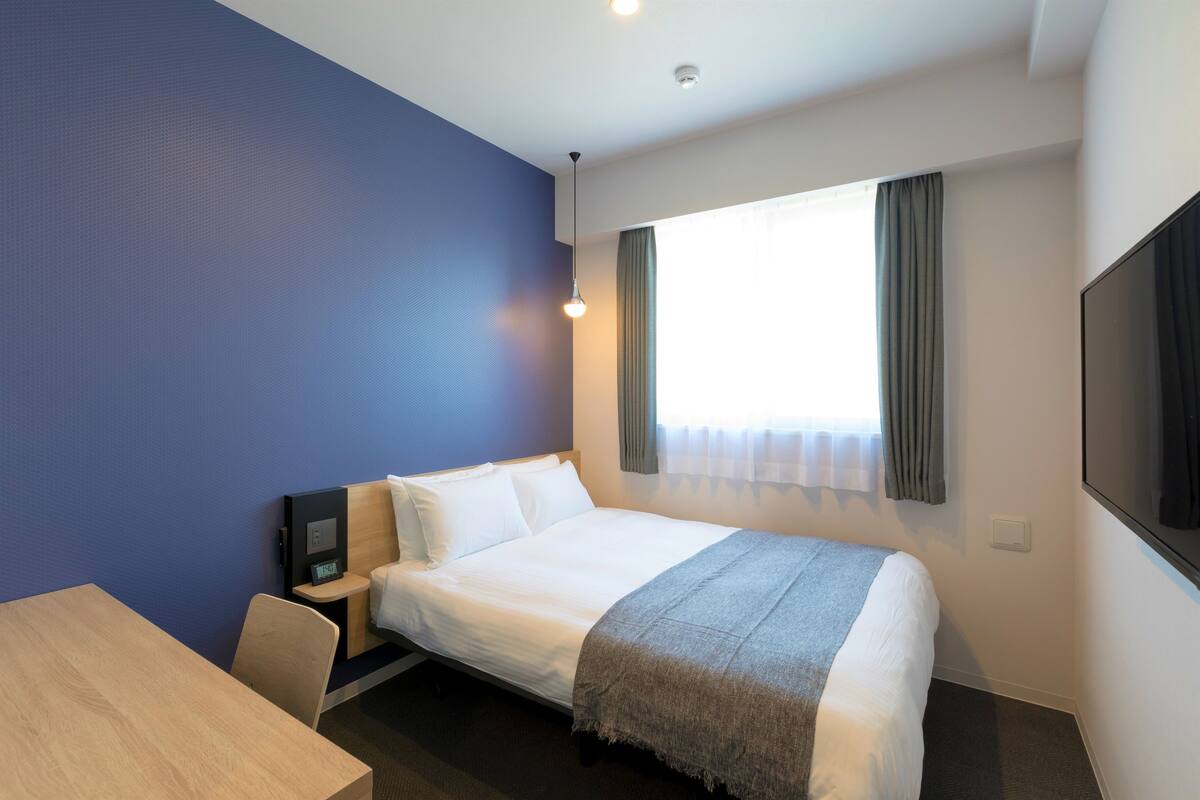 A hotel room with a bed, a desk, a chair, a TV, and a window with curtains.