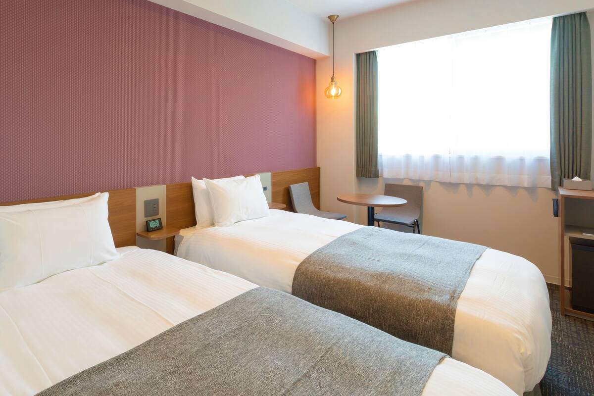 A hotel room with two beds, a small table, a nightstand, and a window with curtains.