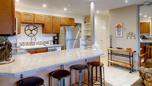 A modern kitchen with wooden cabinets, a central island with stools, a refrigerator, and a coffee maker.
