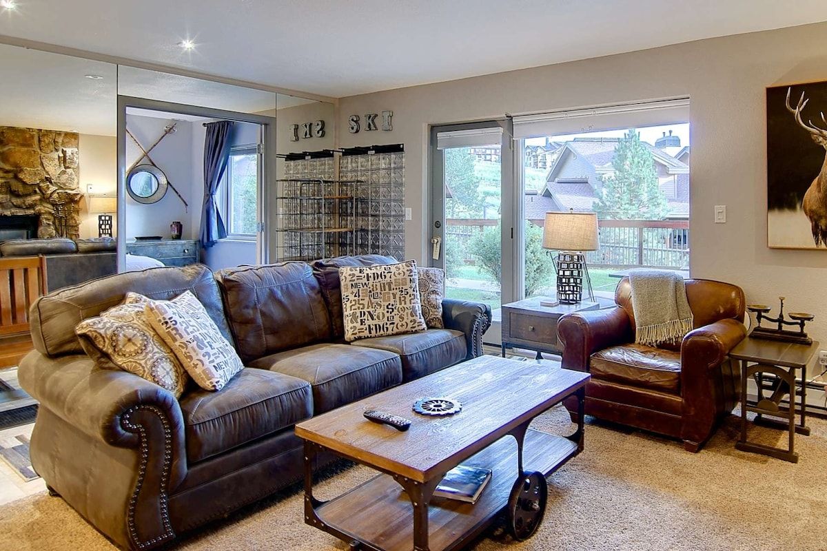 A living room with a brown leather sofa, a wooden coffee table, and a brown leather armchair. There is a bookshelf with books and decorative items, and a stone wall with a fireplace.