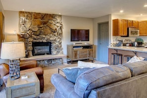 A living room with a stone fireplace, a flat-screen TV, leather sofas, and a kitchen area with wooden cabinets and a microwave. - Ski In & Walk Out-Patio with Slope Views-HotTub (Steamboat Springs)