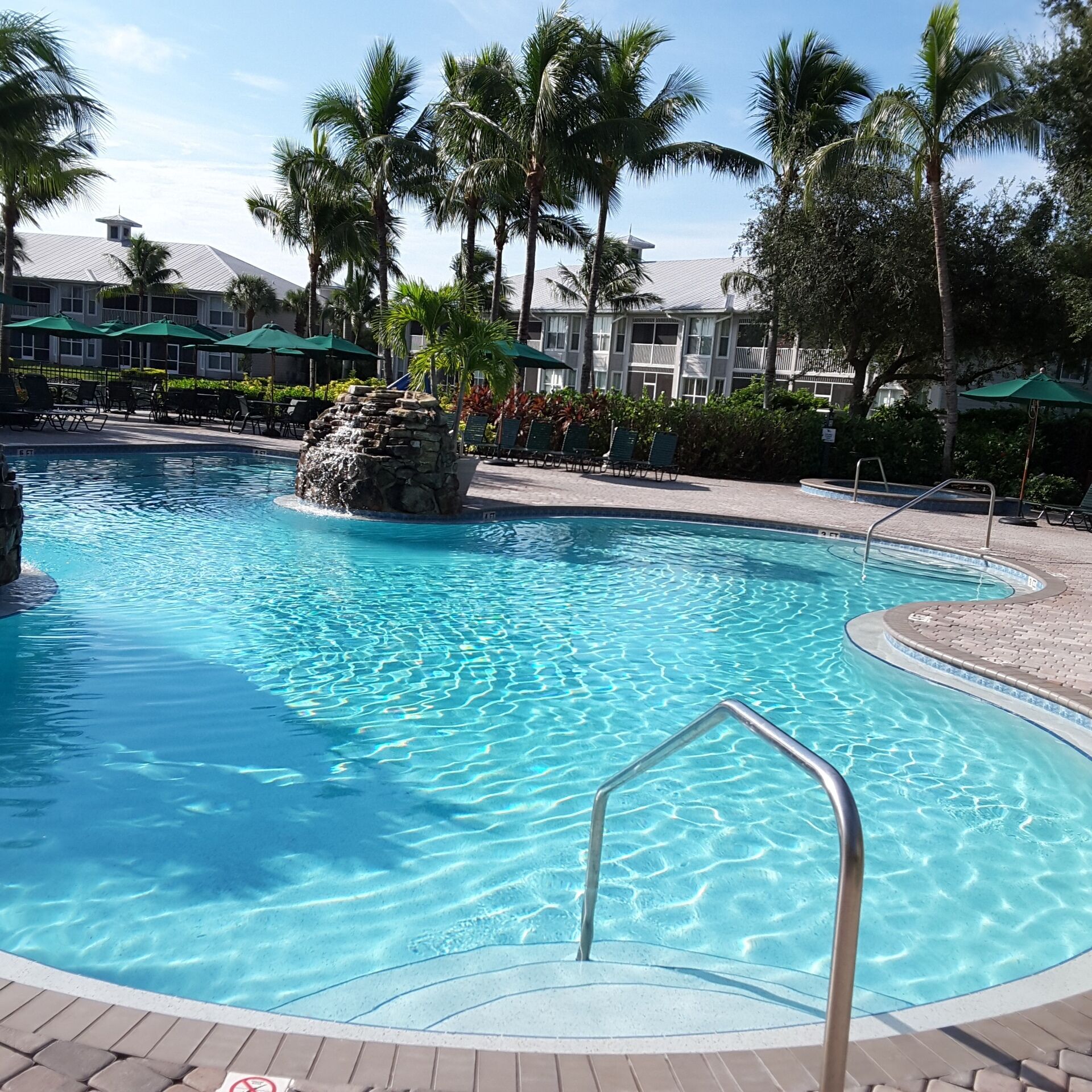 A clear blue pool with a stone waterfall feature, surrounded by lounge chairs and palm trees.