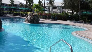 A clear blue pool with a stone waterfall feature, surrounded by lounge chairs and palm trees.