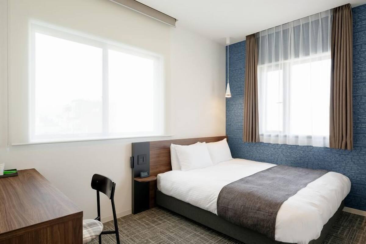 A hotel room with a bed, a desk, a chair, a nightstand, and a window with curtains.