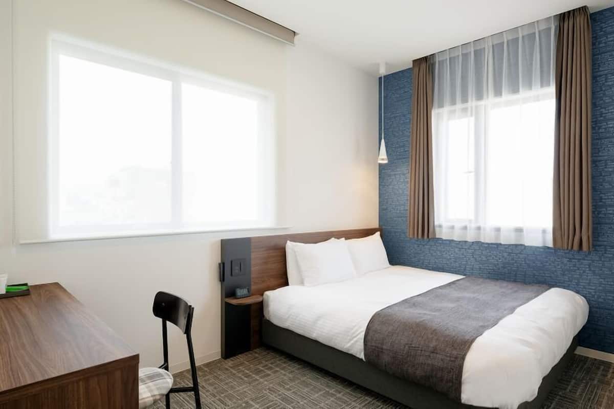 A hotel room with a bed, a desk, a chair, a nightstand, and a window with curtains.