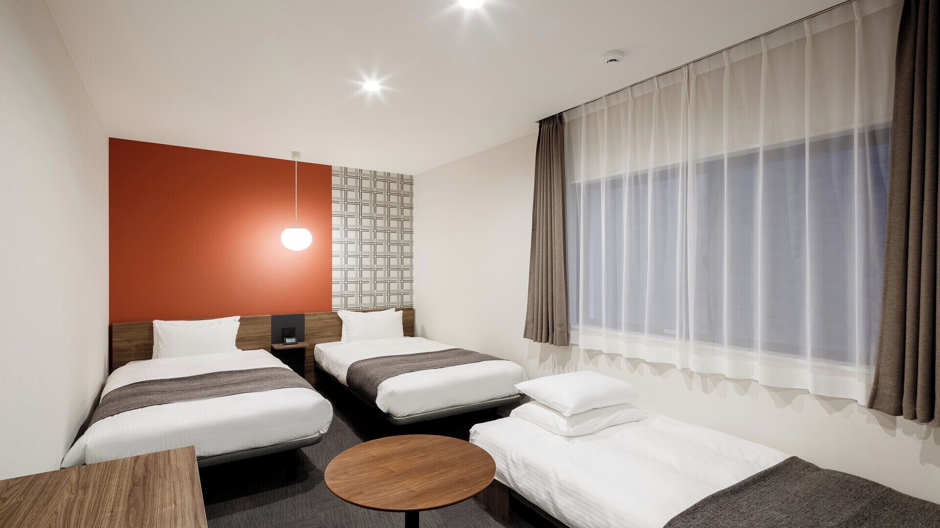 A hotel room with two beds, a small table, a wall-mounted light, and a window with curtains.