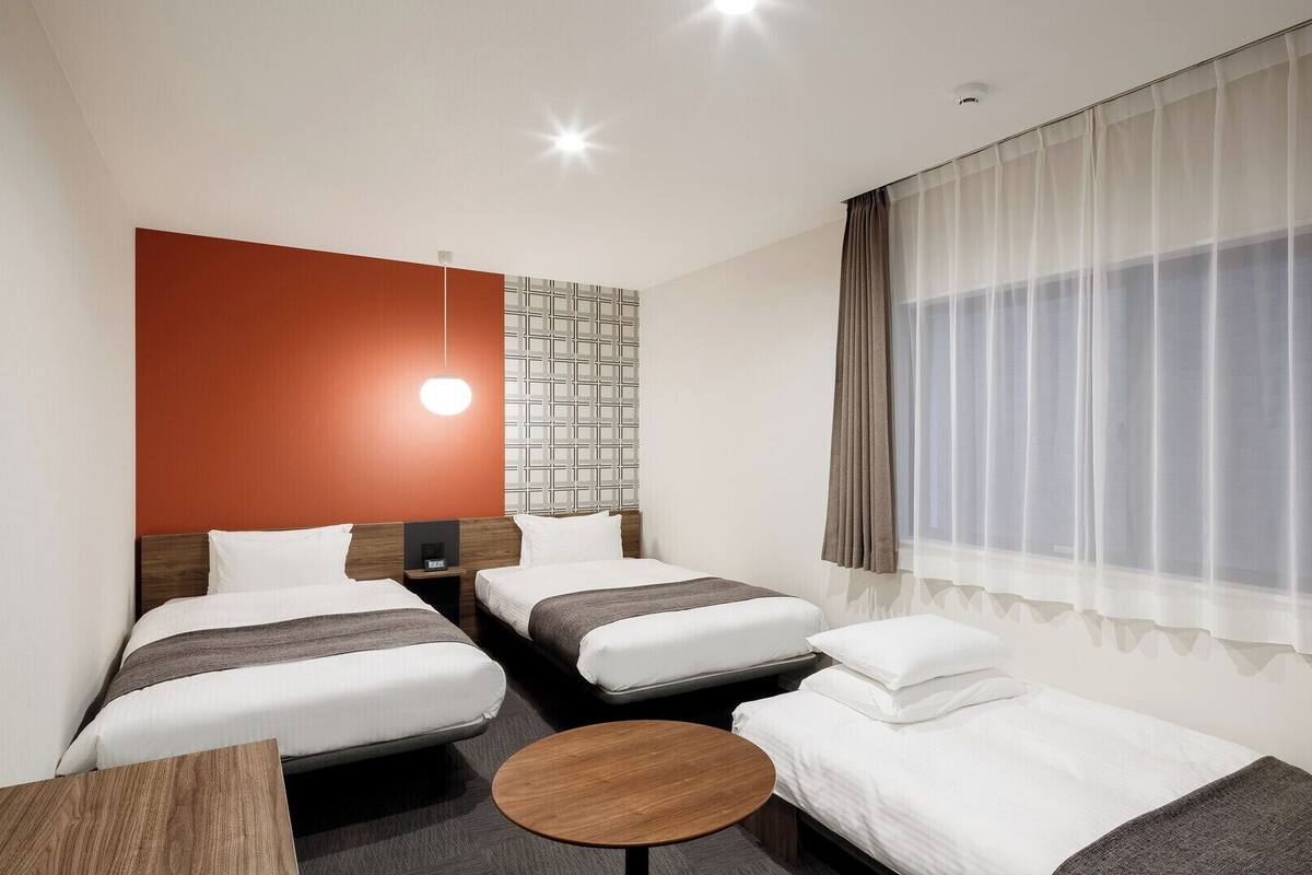 A hotel room with two beds, a small table, a wall-mounted light, and a window with curtains.