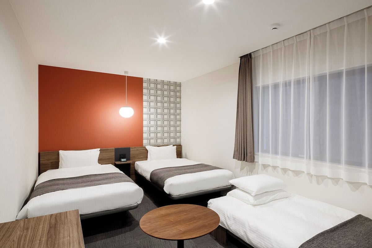 A hotel room with two beds, a small table, a wall-mounted light, and a window with curtains.