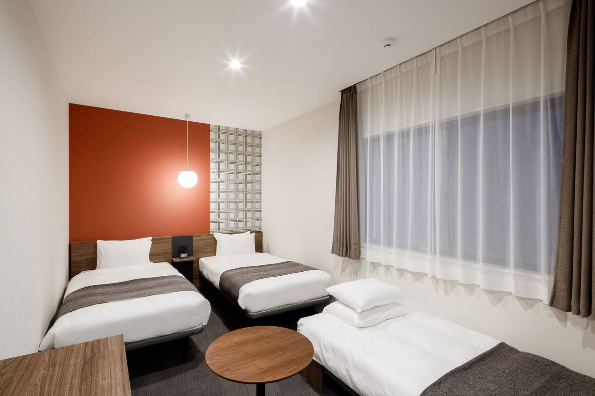 A hotel room with two single beds, a small wooden table, a nightstand, and a large window with sheer curtains.