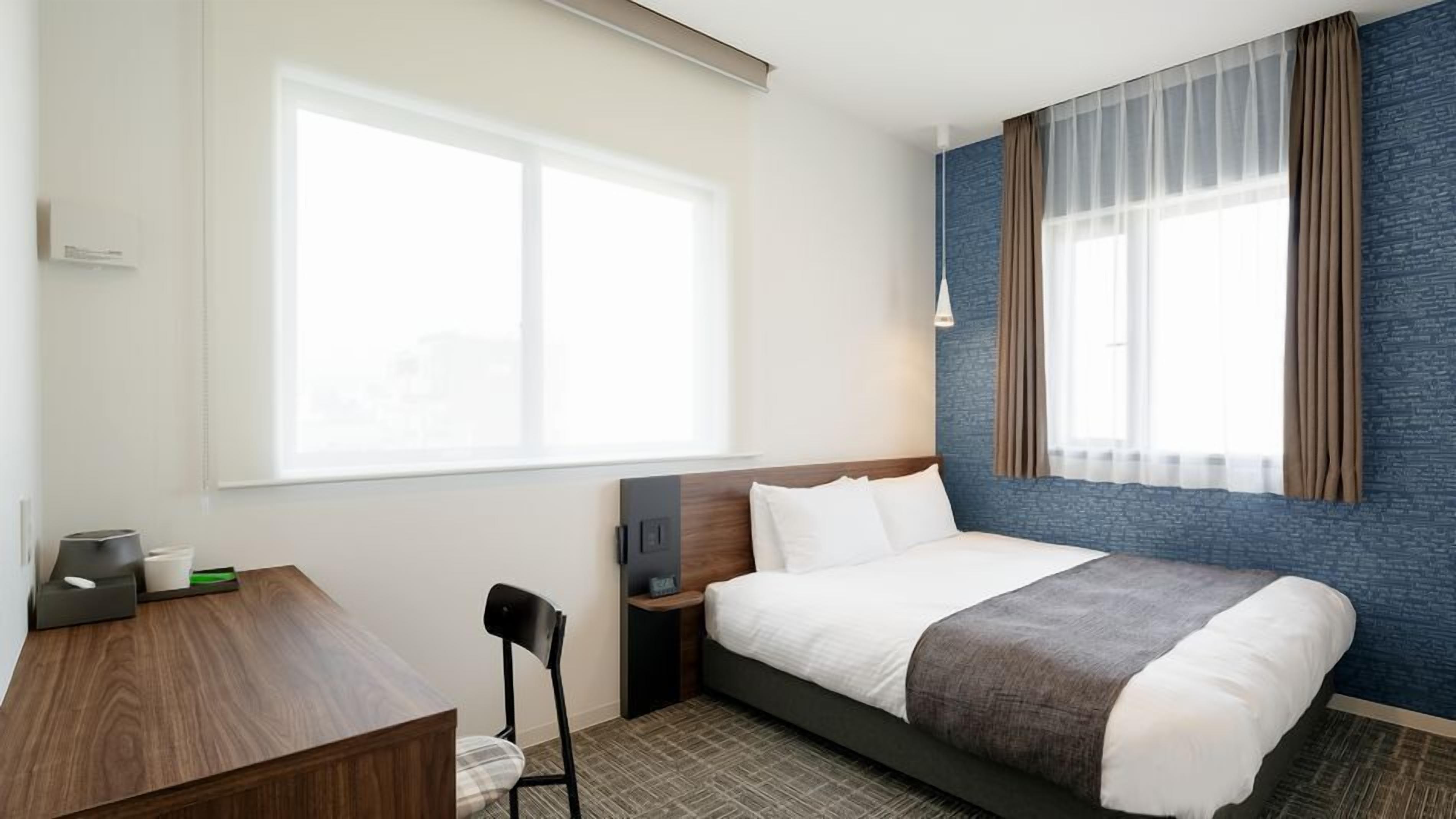 Quarto de hotel com cama, escrivaninha, cadeira, mesa de cabeceira e uma janela grande com cortinas translúcidas.