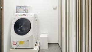 A compact laundry room with a front-loading washing machine, a small white cabinet, and a striped wall. - M’s Hotel Kyoto Station Kizuya (Kyoto)