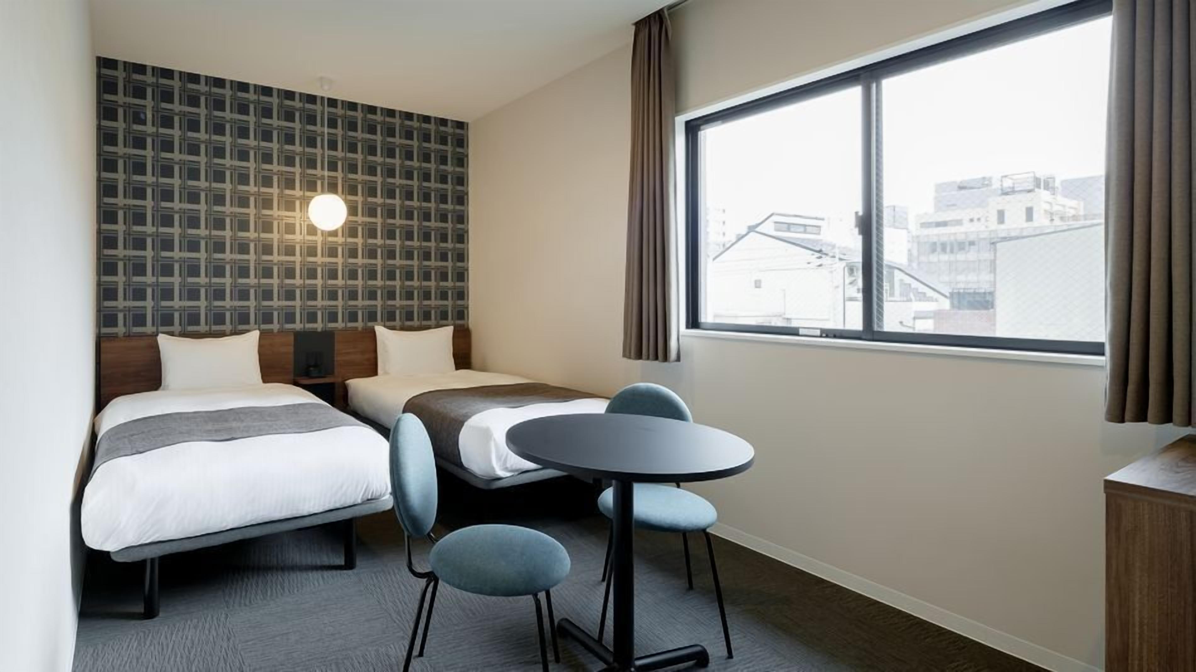 A hotel room with two beds, a small table, and a large window with a city view.