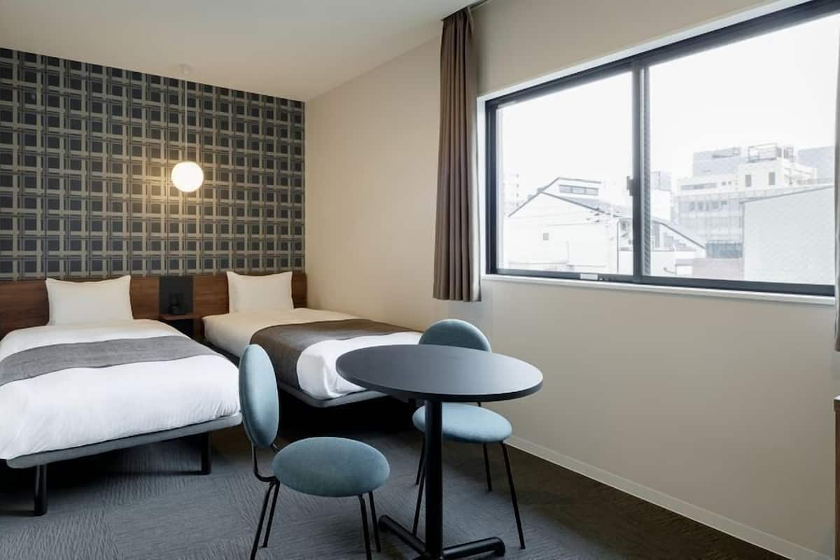 A hotel room with two beds, a small table, and a large window with a city view.