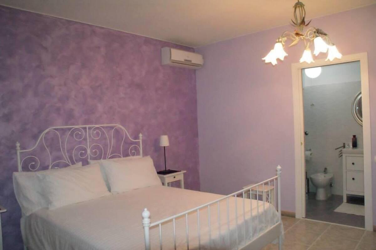 A bedroom with a bed, a nightstand, an air conditioning unit, and a bathroom visible through an open door.