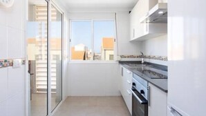 Une cuisine compacte dotée d’une porte-fenêtre coulissante donnant sur un balcon, d’un four encastré et d’une fenêtre qui laisse entrer la lumière naturelle.