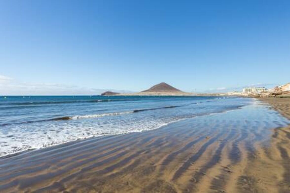 A beach with clear blue water, a sandy shore, and a mountain in the background.