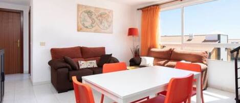 A modern living room with a white dining table and orange chairs, a brown sofa with cushions, and a large window with curtains.
