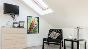 A room with a sloped ceiling, a flat-screen TV mounted on the wall, a wooden dresser, a black chair with a cushion, a patterned rug, and a small table with a decorative item. - Cosy 1 bedroom in Belsize Park (London)