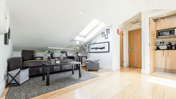 A modern living room with a sofa, coffee table, and a dining area. There is a skylight above the sofa and a kitchen visible through an open door. - Cosy 1 bedroom in Belsize Park (London)