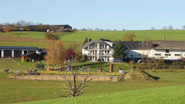 Un cadre rural avec un grand bâtiment blanc doté de nombreuses fenêtres et d’un toit en pente, entouré de champs verdoyants et d’une zone délimitée par une clôture, où se trouve une structure de jeux.