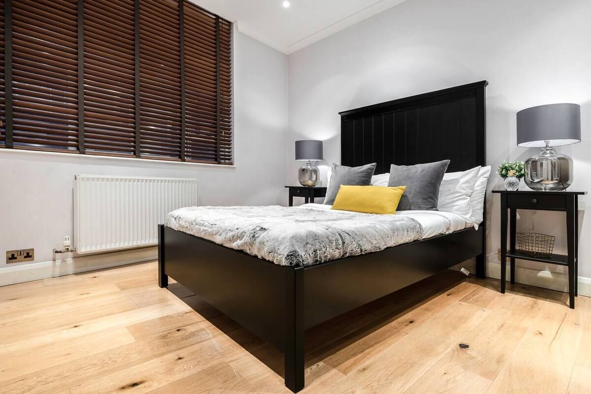 A modern bedroom with a black headboard, a bed with a grey and white patterned bedspread, a yellow accent pillow, and two bedside tables with lamps.