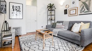 En moderne stue med en grå sofa, et træ sofabord og et mønstret tæppe. Væggene er prydet med indrammede kunstværker, og der står en plante på en hylde.