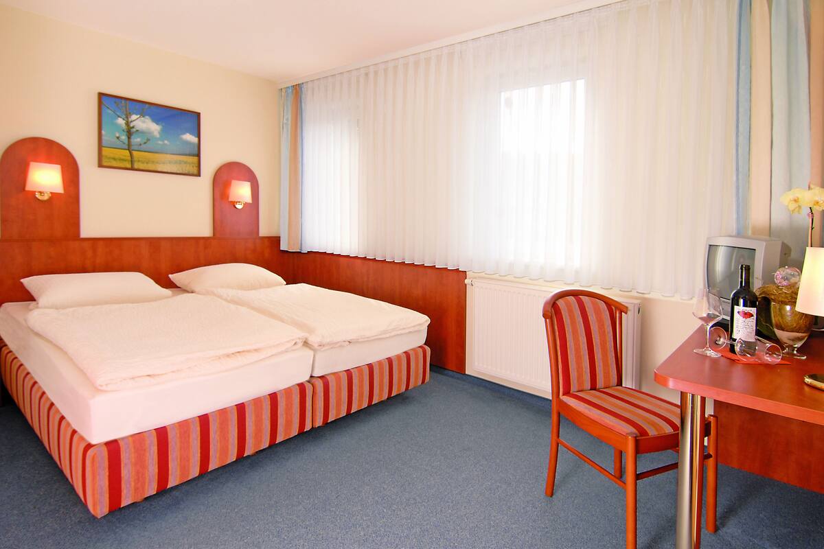 Ein Hotelzimmer mit Doppelbett, einem kleinen Tisch mit Stuhl, einem Fernseher und einem Fenster mit Vorhängen.