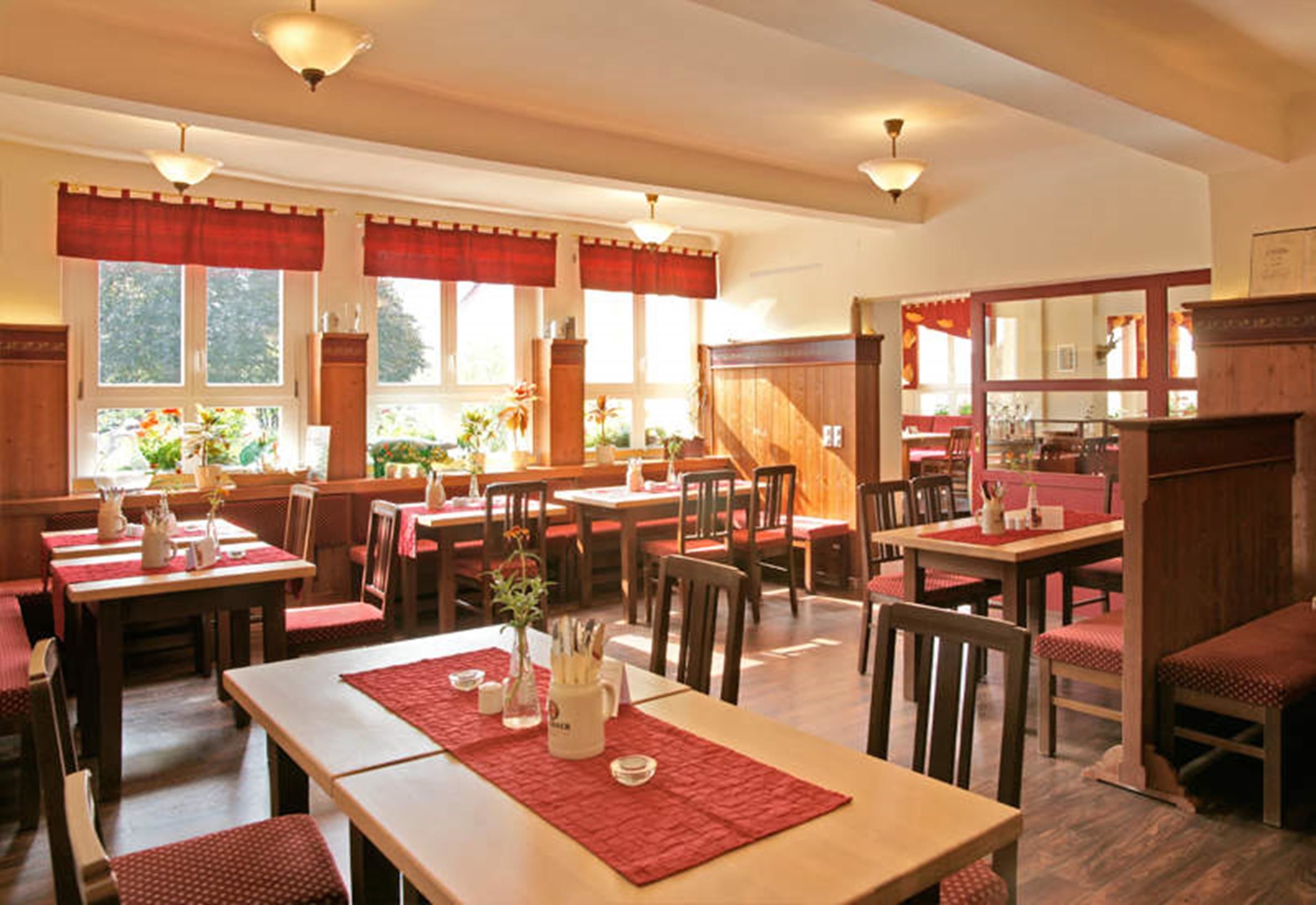 A restaurant with wooden tables and chairs, red tablecloths, and large windows with curtains.