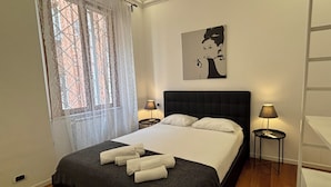 Une chambre à coucher avec un lit, deux lampes de chevet, une fenêtre ornée de rideaux en dentelle et une œuvre d’art accrochée au mur.