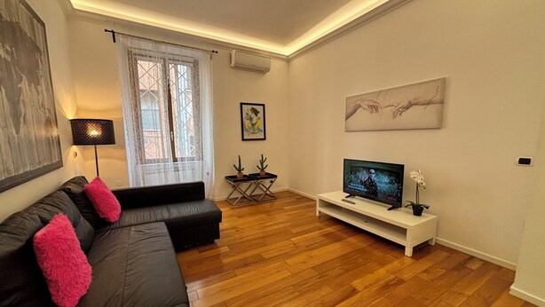 Un salon avec un canapé noir, un téléviseur posé sur un meuble blanc, une fenêtre ornée de rideaux légers et un tableau accroché au mur.
