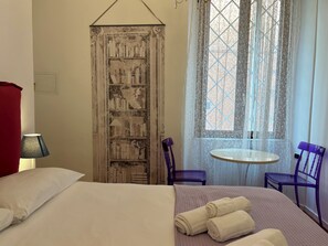 Habitación de hotel con cama, una mesita, dos sillas, ventana con cortinas y un panel decorativo en la pared.