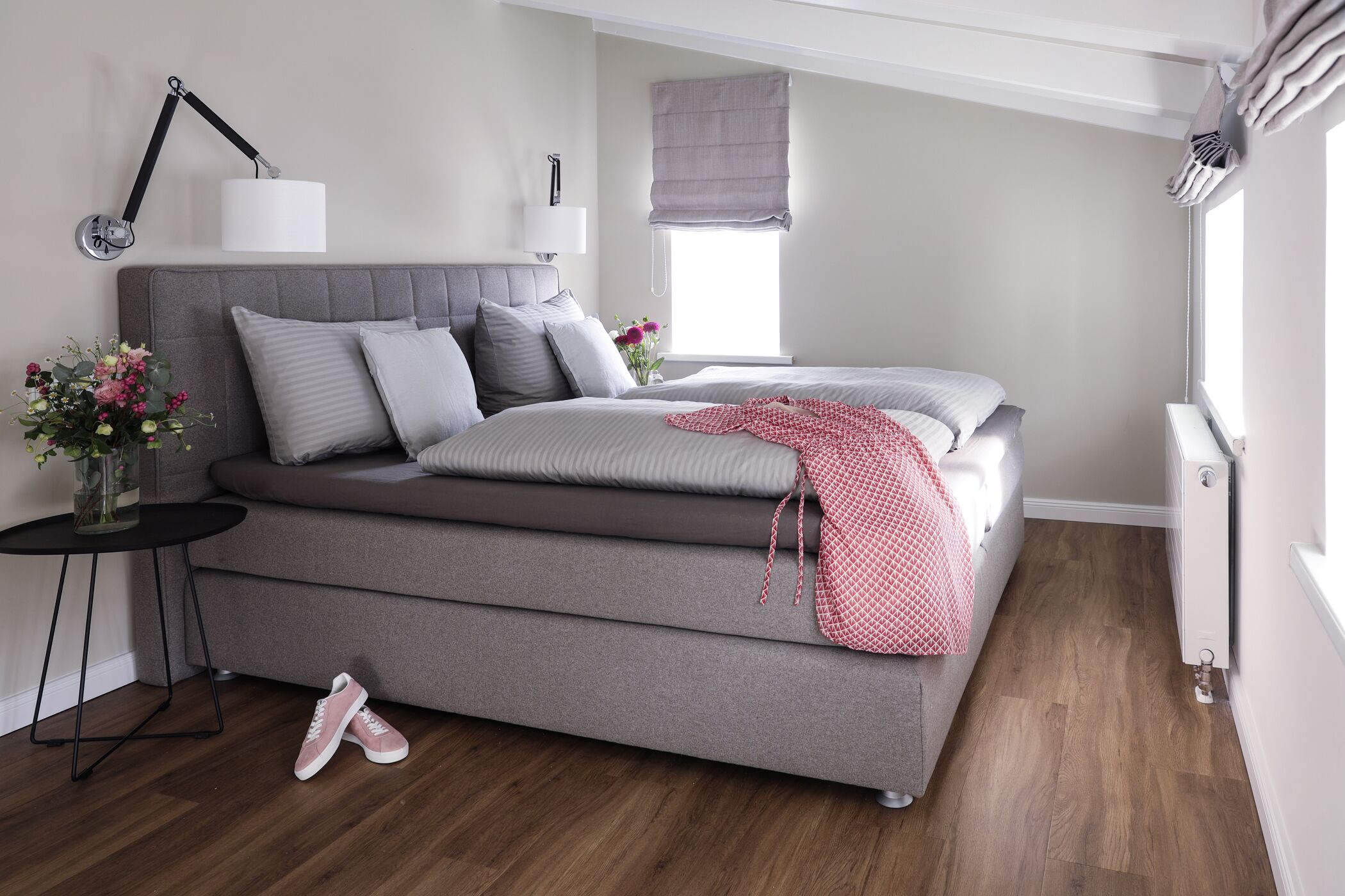 A modern bedroom with a grey sofa bed, a bedside table with a lamp, a window with a blind, and a small table with a vase of flowers.