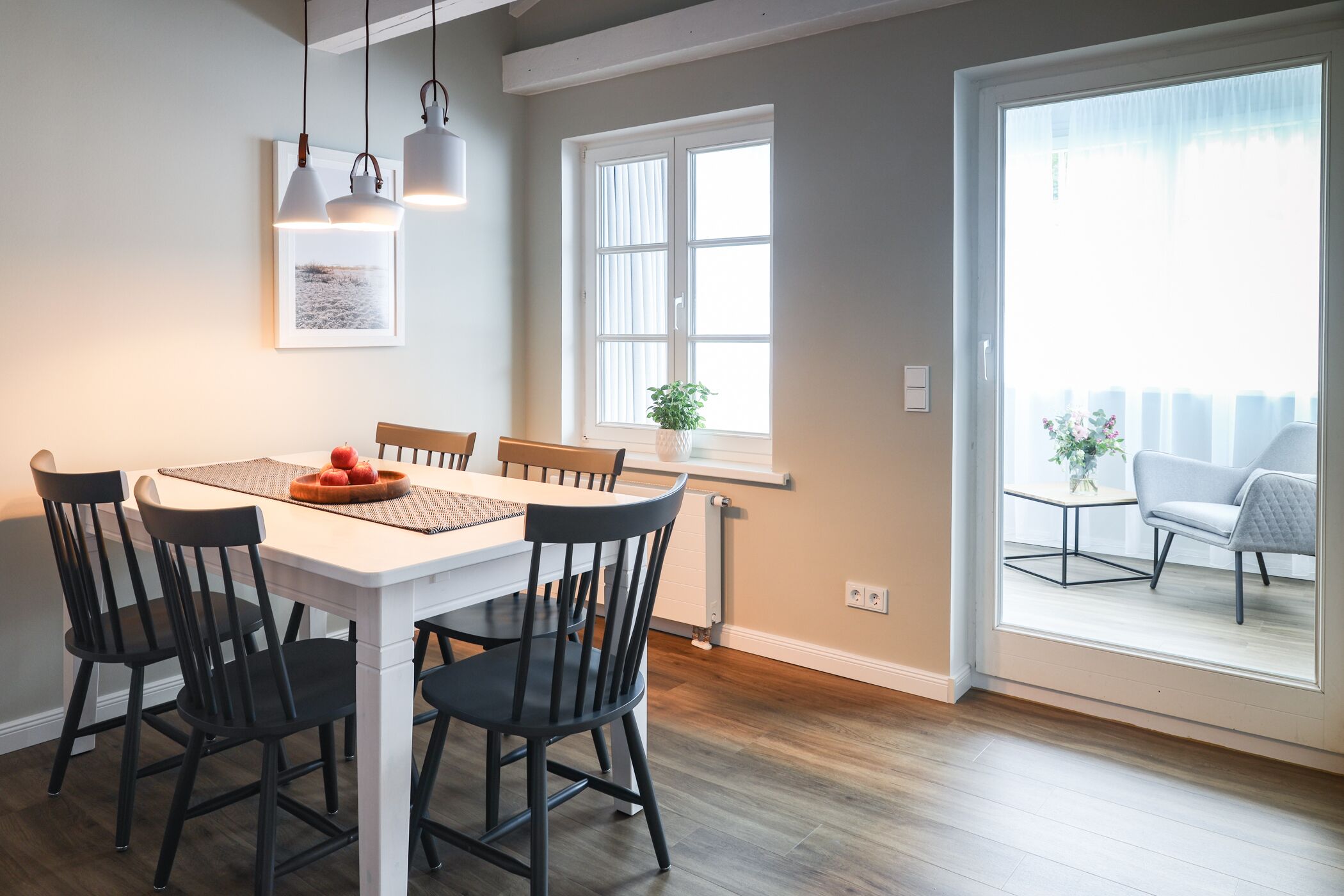 A dining area with a white table, black chairs, and pendant lights. There is a window with a view of a balcony.