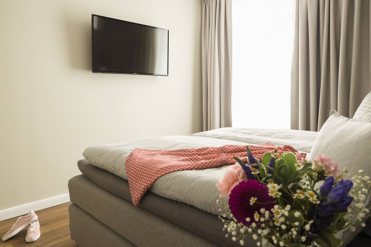 A bedroom with a bed, a TV mounted on the wall, and a window with curtains.