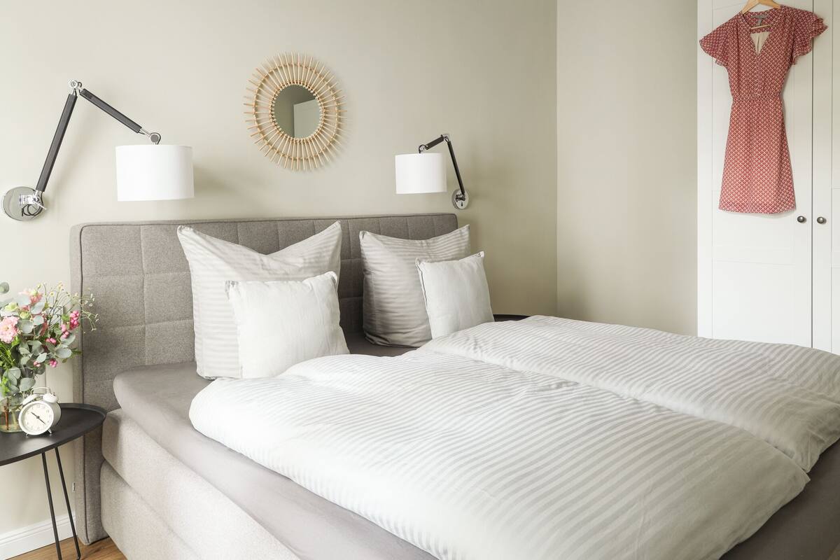 A neatly made bed with white bedding, two bedside lamps, a wall clock, and a mirror.