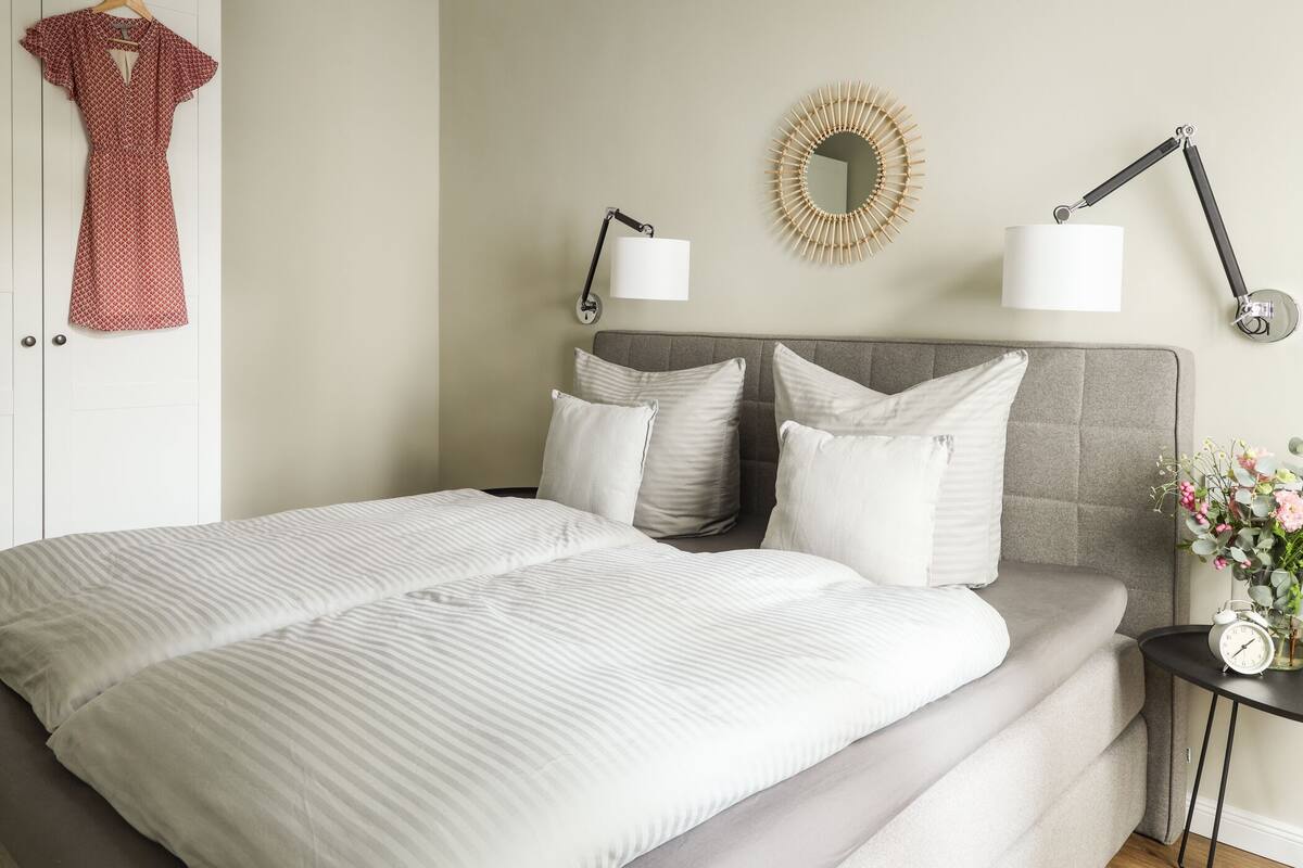 A neatly made bed with a grey headboard, two bedside lamps, and a mirror on the wall.