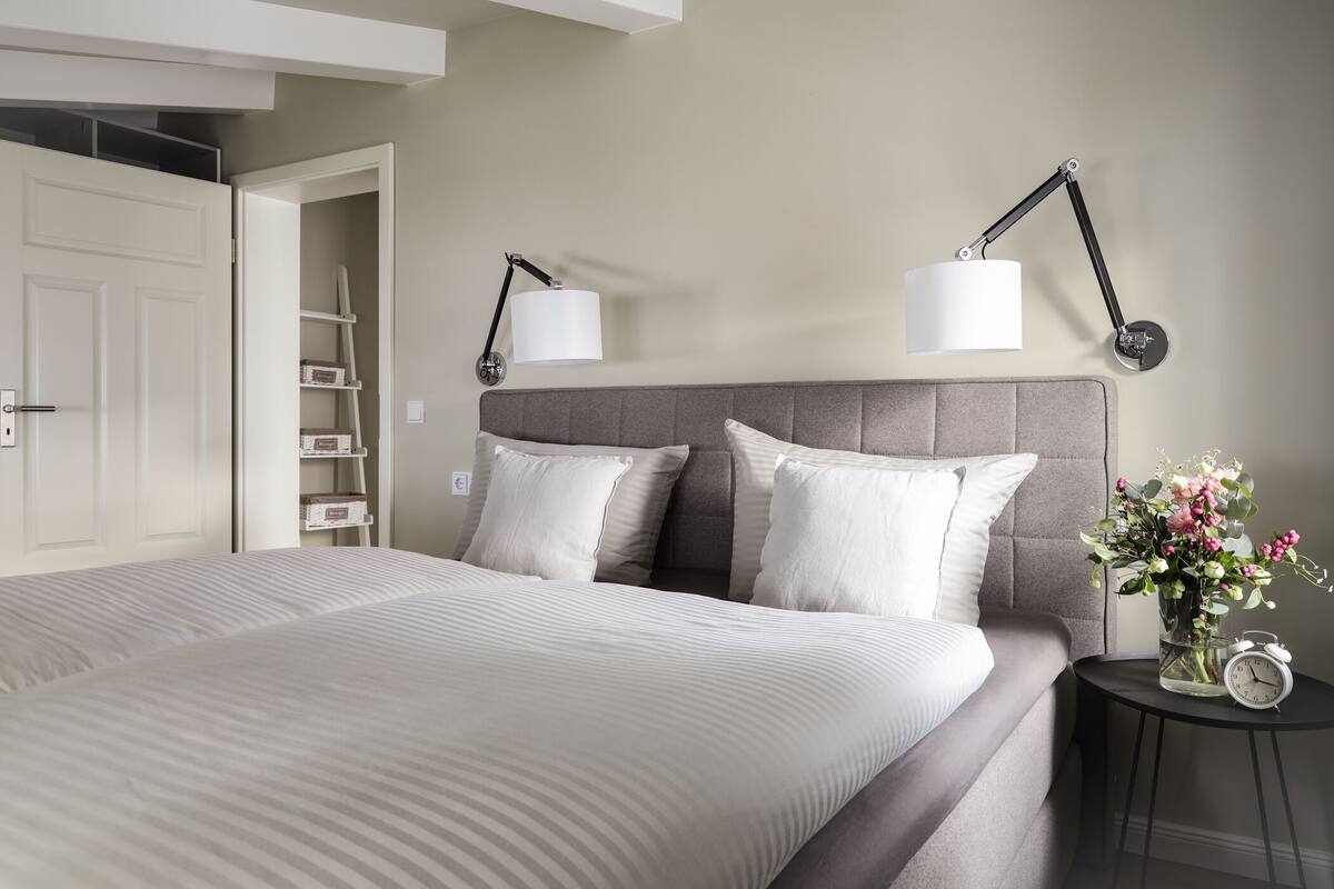 A neatly arranged bedroom with a large bed, two bedside lamps, a small table with a vase of flowers, and a clock.