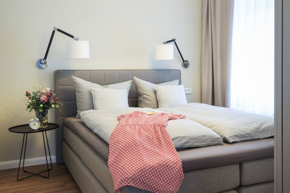 A modern bedroom with a bed, two bedside lamps, a small table with a clock, and a vase with flowers.