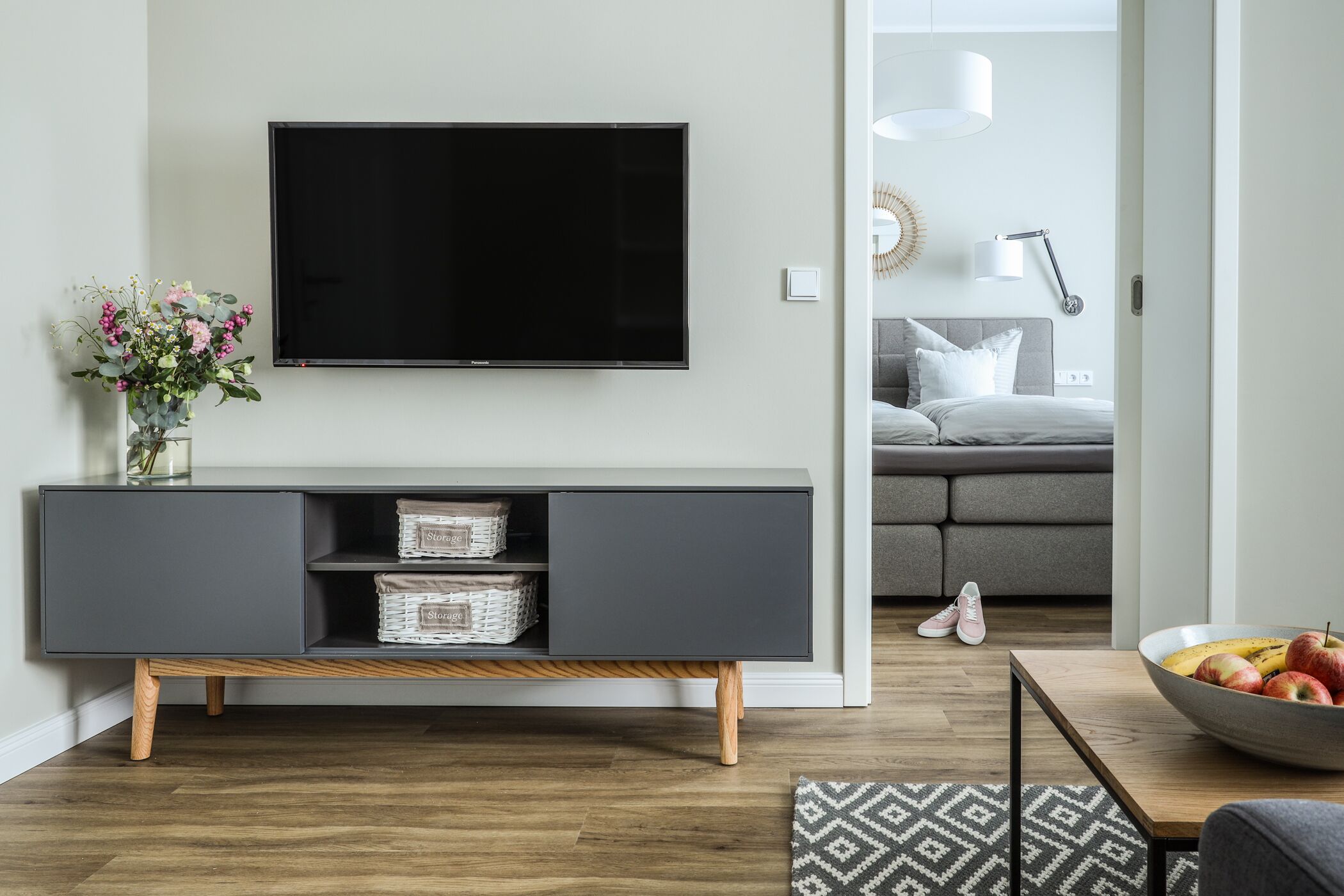A modern living room with a flat-screen TV mounted on the wall, a grey console table with wicker baskets, a patterned rug, and a bowl of fruit on a side table.