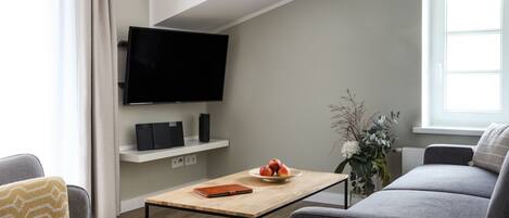 A modern living room with a flat-screen TV, a coffee table, a sofa, and a small table with a bowl of fruit.