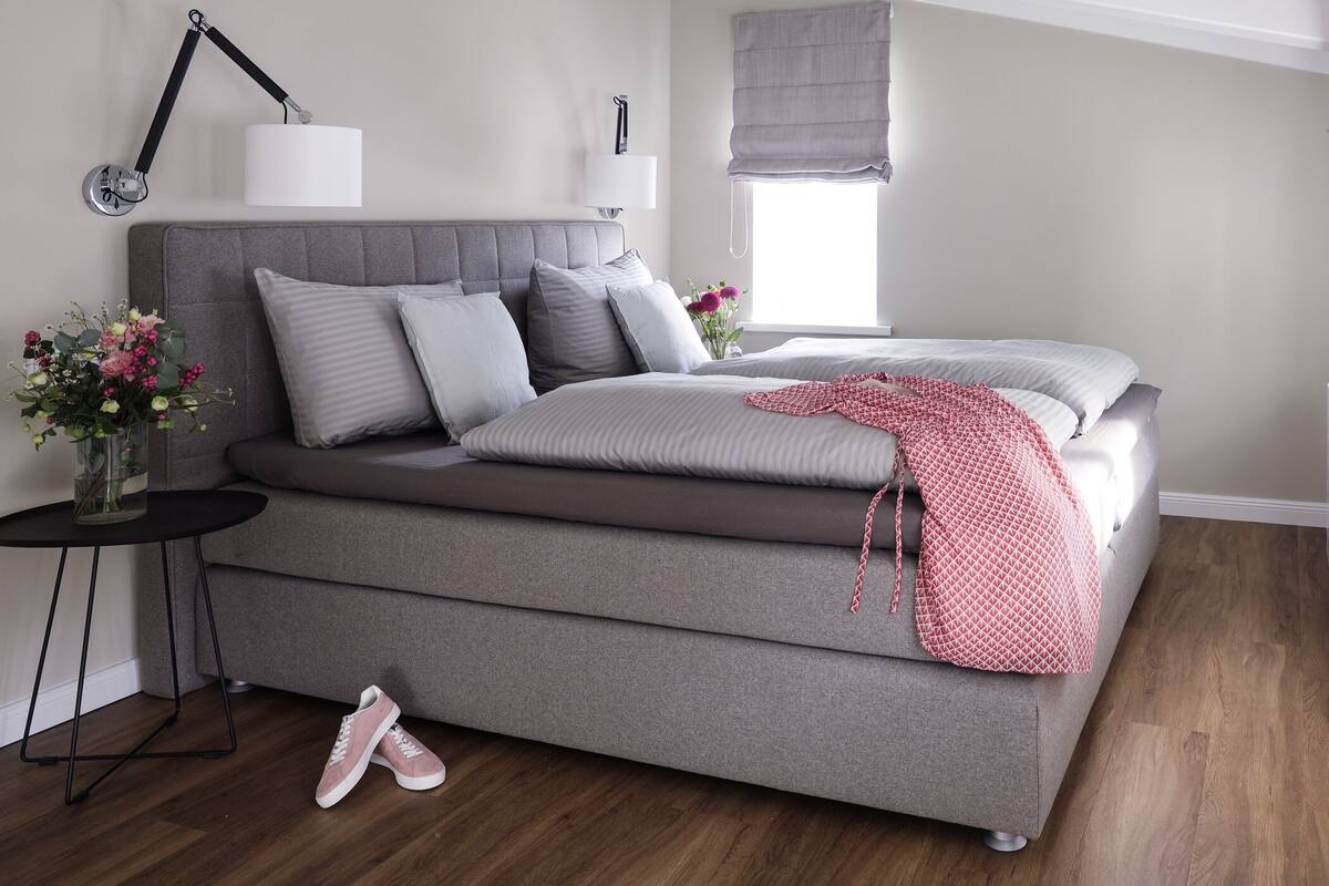 A modern bedroom with a grey sofa bed, a small side table with a vase of flowers, and a pair of shoes on the floor.