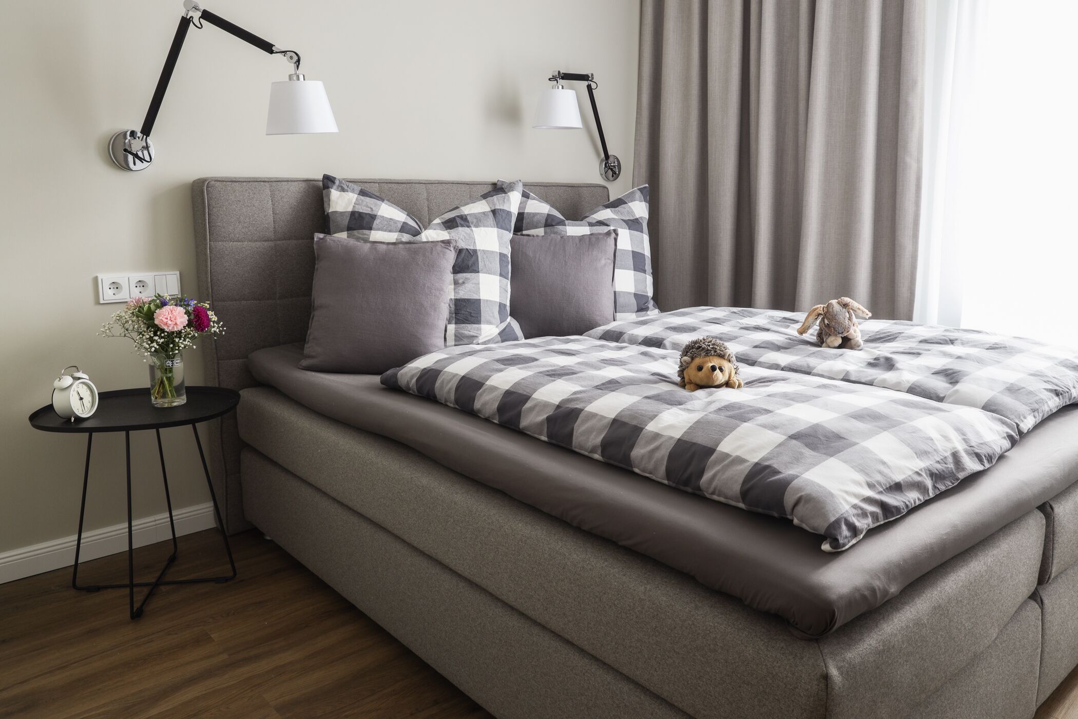 Een netjes opgemaakt slaapkamer met een bed, twee wandlampen, een klein nachtkastje met een klok en bloemen, en een knuffel op het bed.