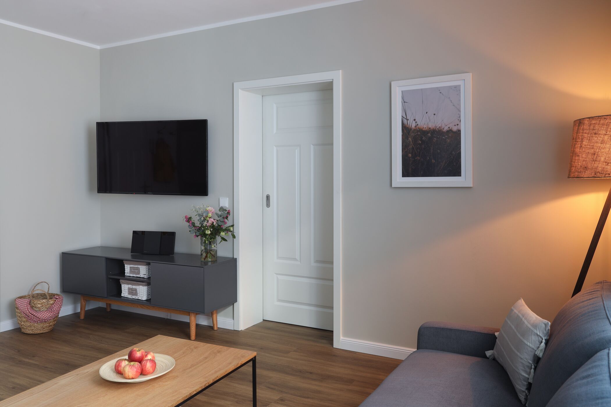 A modern living room with a flat-screen TV, grey sofa, and a wooden coffee table with apples.