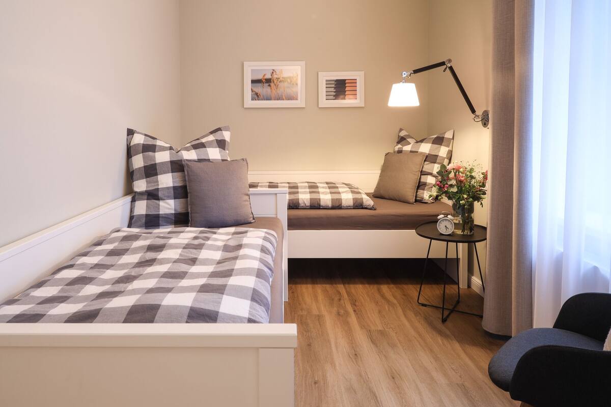 A compact hotel room with two beds, a small table, a chair, and a lamp.