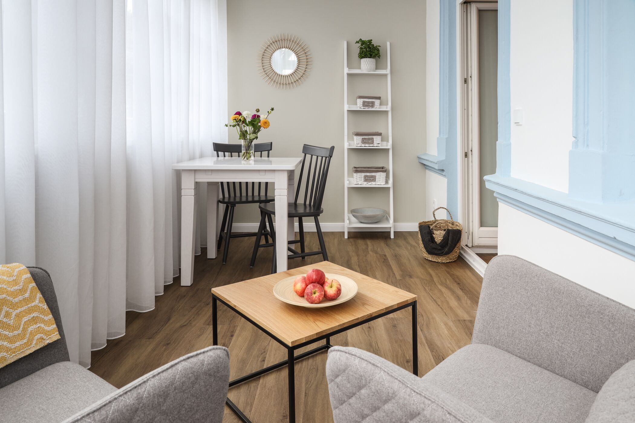 Ein modernes Wohnzimmer mit einem Holztisch, zwei grauen Sofas, einem weißen Esstisch mit Stühlen, einer weißen Schrankwand und einem Korb auf dem Boden.
