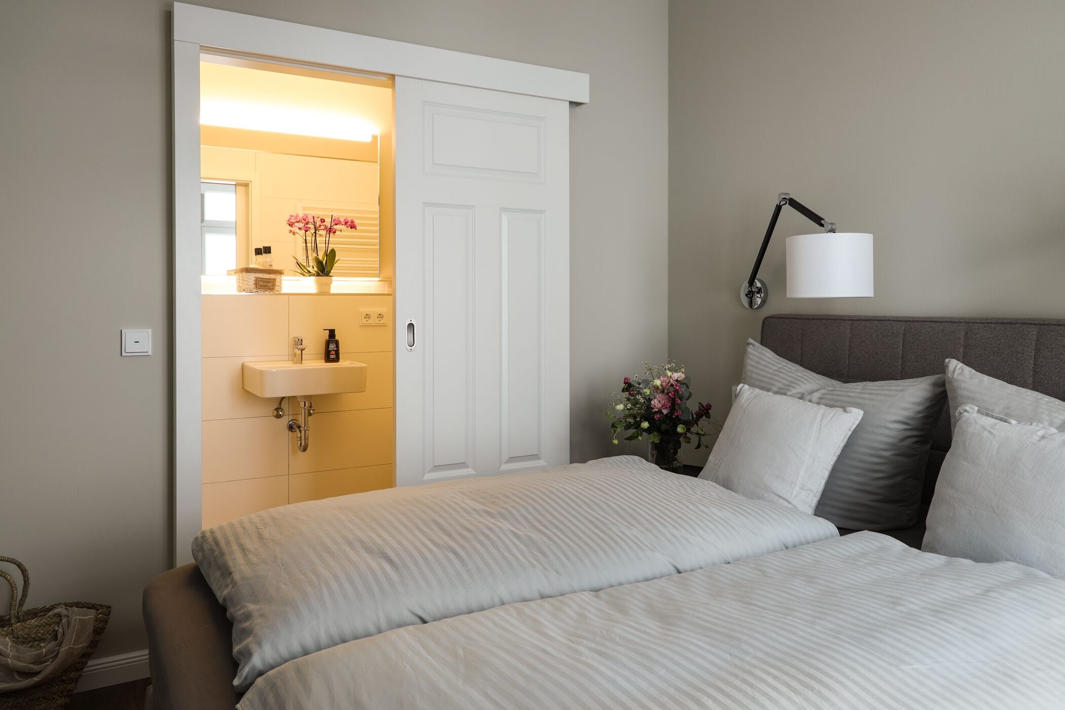 Une chambre à coucher comprenant un lit, une table de chevet, une lampe, une salle de bain avec un lavabo et une porte donnant sur une autre pièce.