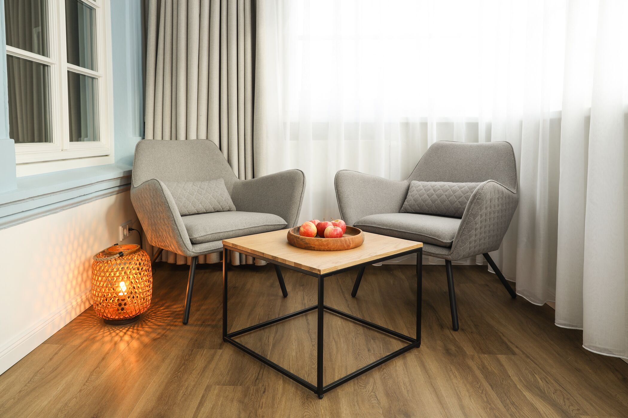 A modern living room with two grey armchairs, a wooden coffee table with a fruit bowl, a patterned floor lamp, and a window with sheer curtains.