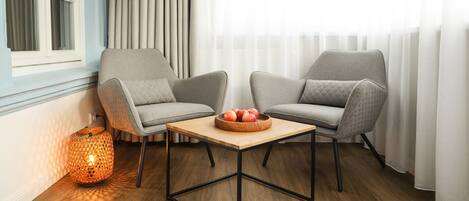 A modern living room with two grey armchairs, a wooden coffee table with a fruit bowl, a patterned floor lamp, and a window with sheer curtains.